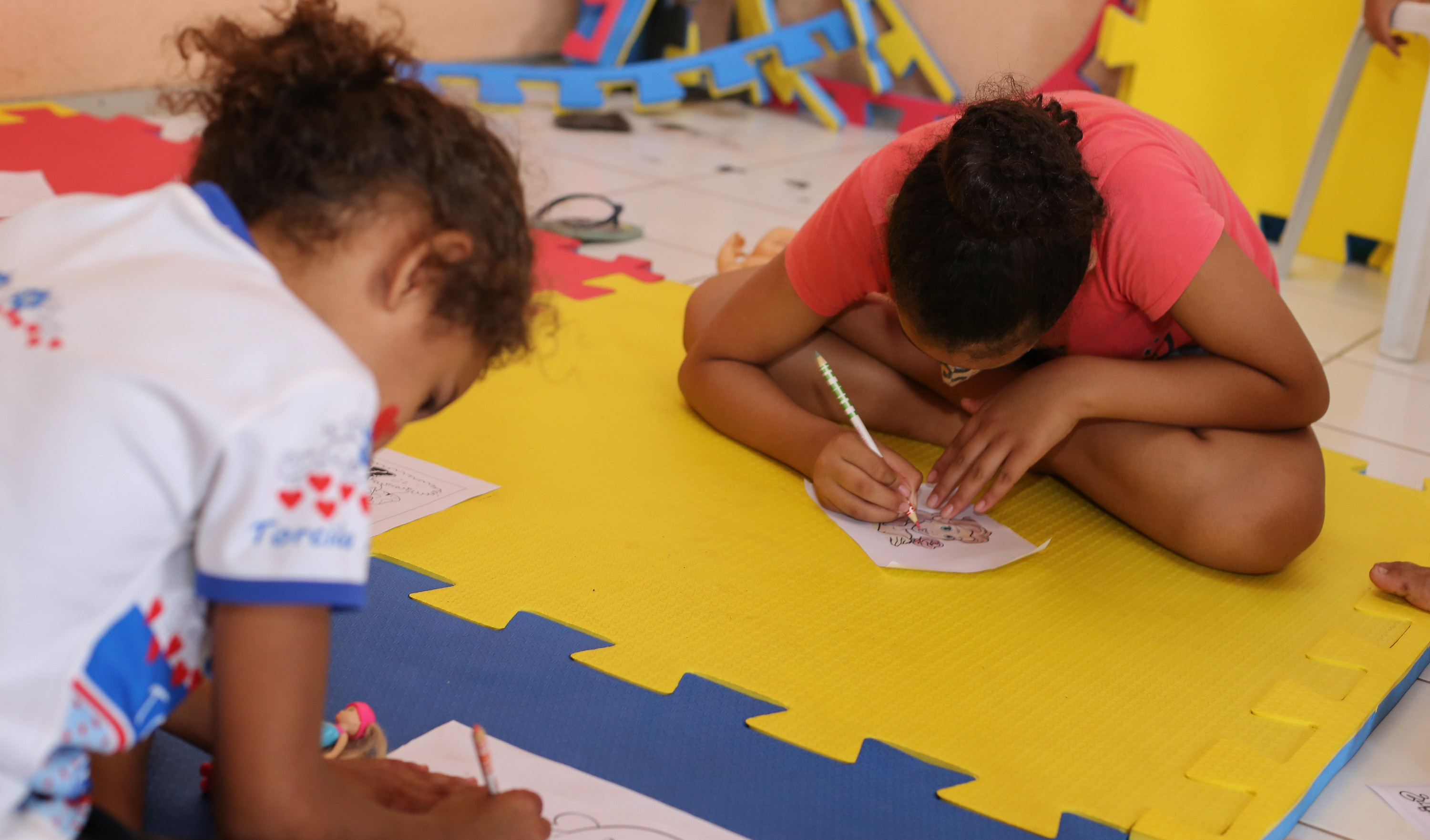 duas crianças, de máscara, pintam um desenho, sentadas sobre um tapete de brincadeiras colorido de amarelo, azul e vermelho